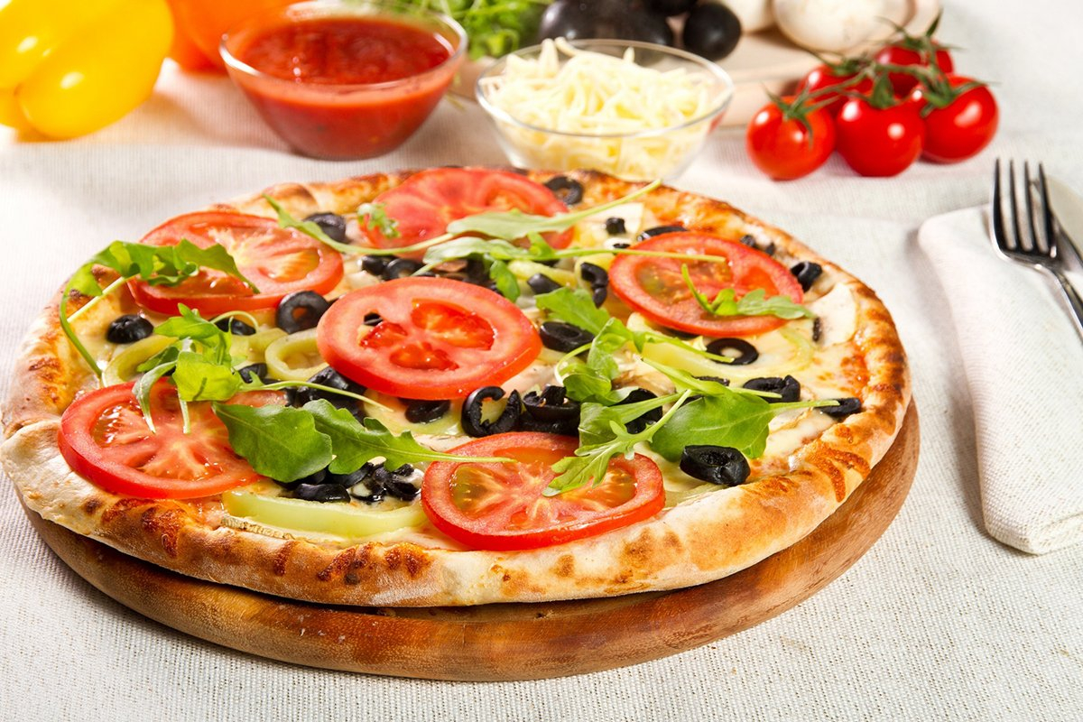 على طبق خشبي تظهر بيتزا مغطاة بمختلف انواع الخضار كالبندورة والفليفله ز بازراق خضراء اخلى و بجانبها حبات من البندورة و صحن من الجبنة وصوص البدورة و حبة فليفه و غيرشها 